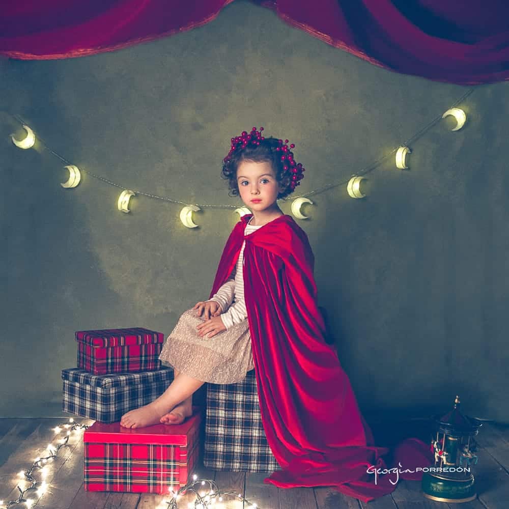 nena amb llumetes al fons i capa i accessoris vermells sentada a sobre de regals sessio estudi infantil de Nadal a Granollers